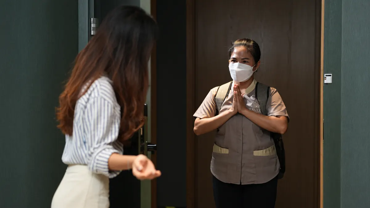 Terapis Truly Home Massage berseragam dan bermasker menyapa pelanggan dengan salam hormat di depan pintu saat tiba di rumah